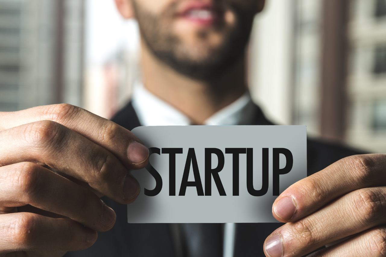 Zbliżenie na dłonie uśmiechniętego mężczyzny w eleganckim garniturze, który trzyma przed sobą szarą kartę z dużym, czarnym napisem STARTUP. Zdjęcie symbolizuje rozwój nowej firmy oraz wsparcie biznesowe, jakim jest profesjonalna obsługa prawna startupów.